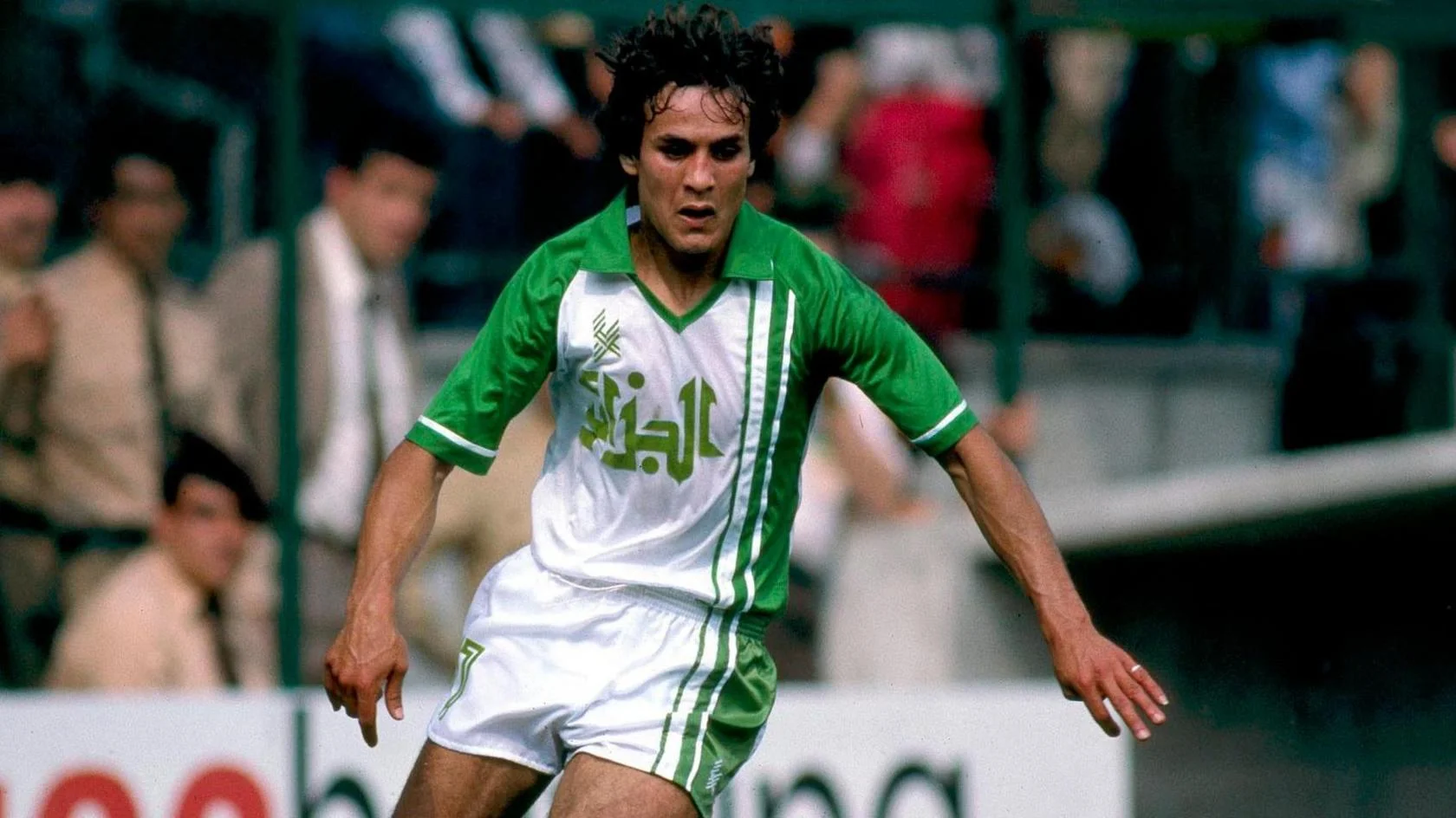 Algeria's Rabeh Madjer seen in action at the 1982 World Cup in Spain. He is wearing predominantly white shorts with green vertical trim on one side and a matching shirt which sees the same green lines carry on up from the shorts onto Madjer's torso to meet a large green collar and green sleeves. Written on the chest, also in green, is the name of Algeria in Arabic script. Madjer is looking down at a ball which is out of shot and leaning to his left, with arms slightly spread for balance, as he looks to turn. His dark hair is bouncing slightly as he moves. Blurred in the background are members of the crowd, including what appears to be some members of stadium security dressed in khaki colours