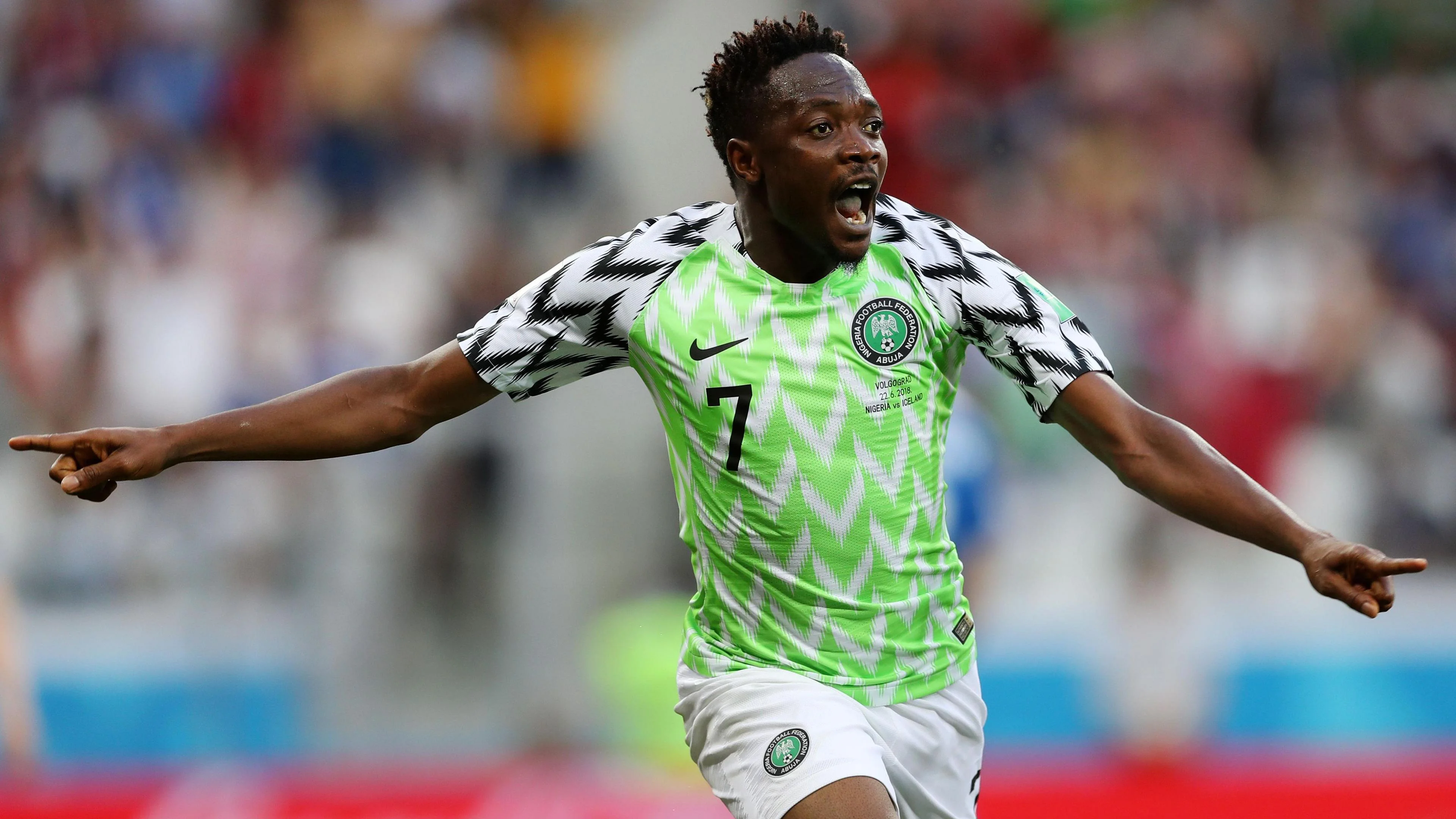 Ahmed Musa in the neon green Nigeria home shirt worn at the 2018 World Cup in Russia. The design includes white chevrons on the green chest and black chevrons on the white sleeves. Musa, who is visible from the thighs up, has his arms spread wide and his mouth open to scream in celebration as he runs after scoring a goal. A packed crowd is blurred in the background