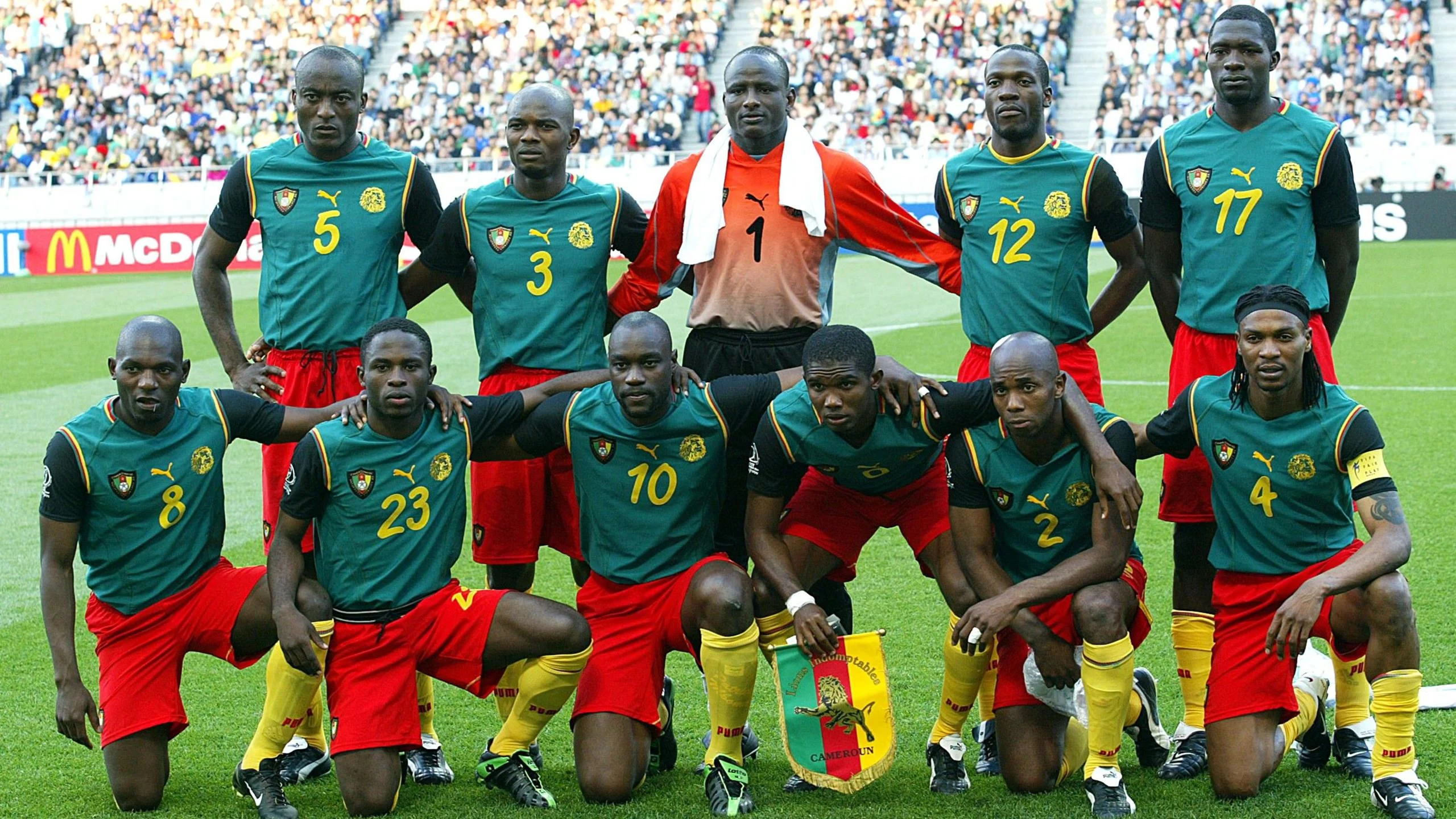 A Cameroon team photograph taken on the pitch before a game at the 2002 World Cup. Six players are down on one knee or on their haunches in the front row, with captain Samuel Eto'o, third from the right, holding a team pennant. The goalkeeper is in the centre of five players standing in the back row and has a white towel draped around his neck. The outfield players wear a kit made up of yellow socks, red shorts and a green sleeveless shirt which has had black upper-arm sleeves sewn on to it. The goalkeeper sports an orange top and black shorts 